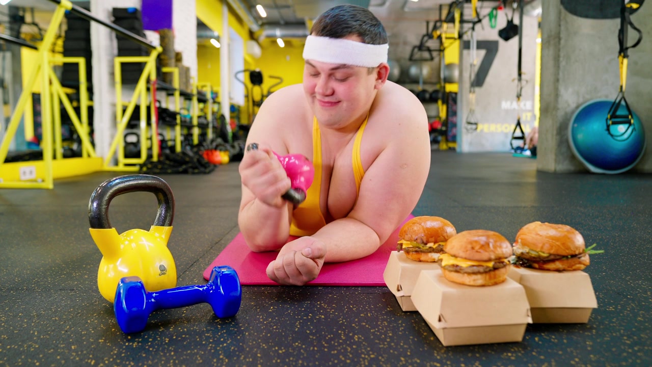Uomo in sovrappeso che fatica con un kettlebell mentre è tentato dagli hamburger in palestra