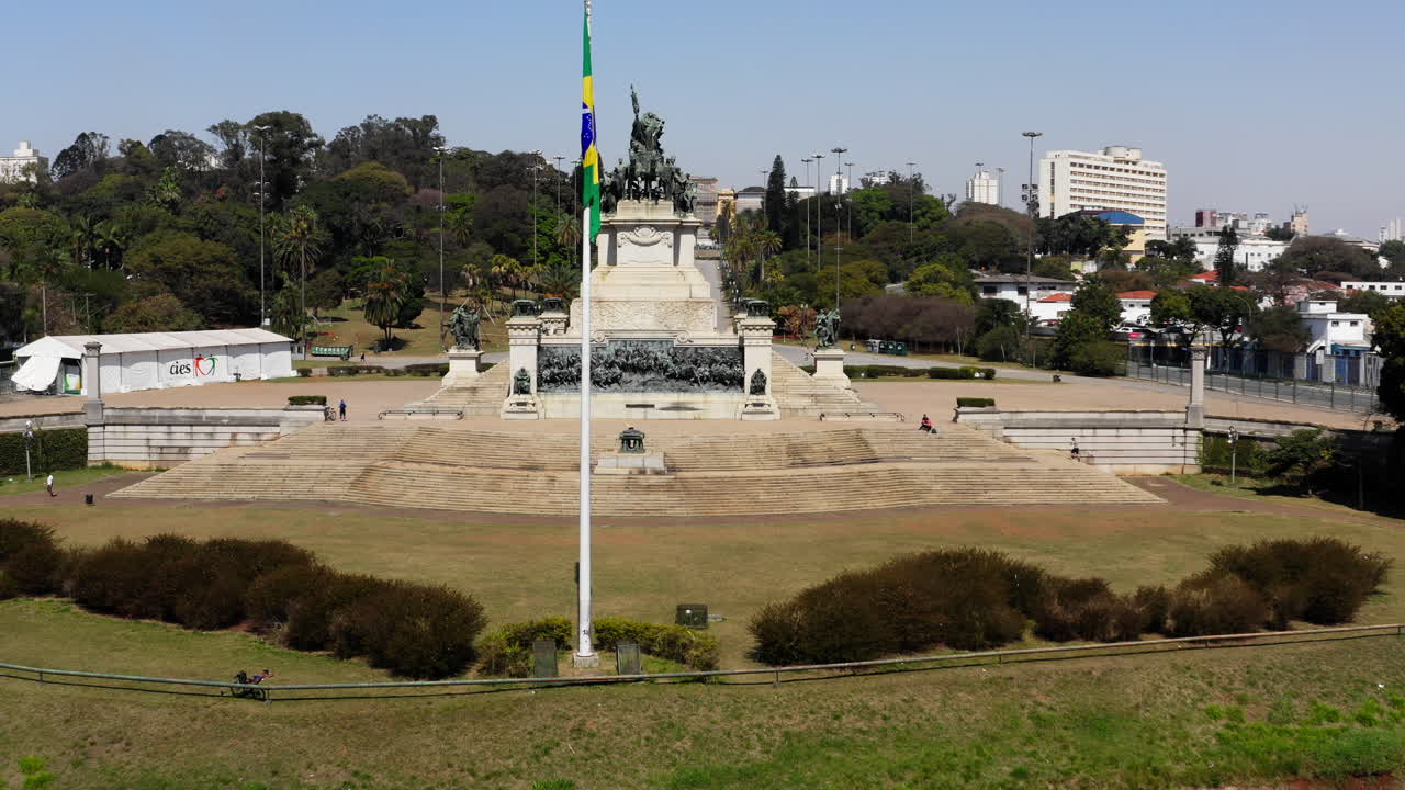 Ripresa aerea in avvicinamento al Monumento all'Indipendenza che passa attraverso la bandiera brasiliana in primo piano, con il Museo dell'Ipiranga sullo sfondo della scena.