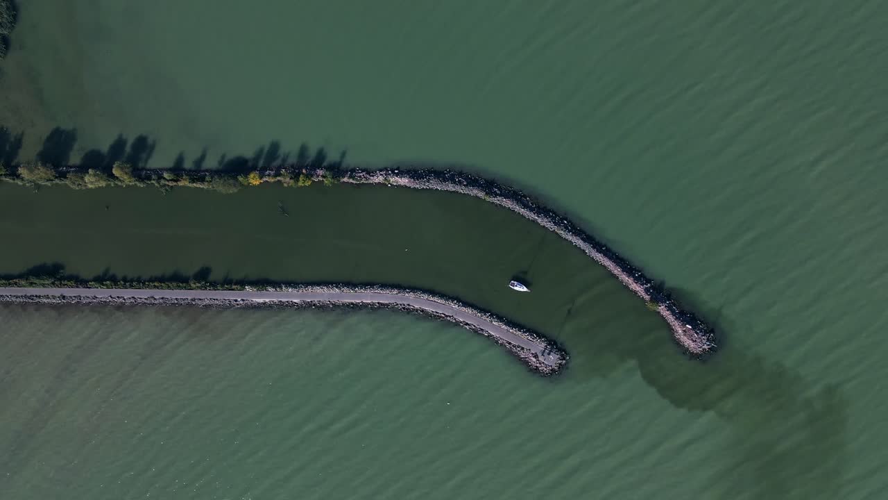 An aerial footage of a small boat docked in a narrow waterway protected by two curved piers extending into a greenish lake.