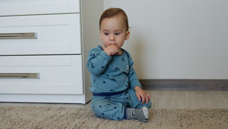 sweetest toddler in blue pajama sitting on the floor of the room beside the chest of drawers. baby boy staring at one point holding a finger in his mouth.