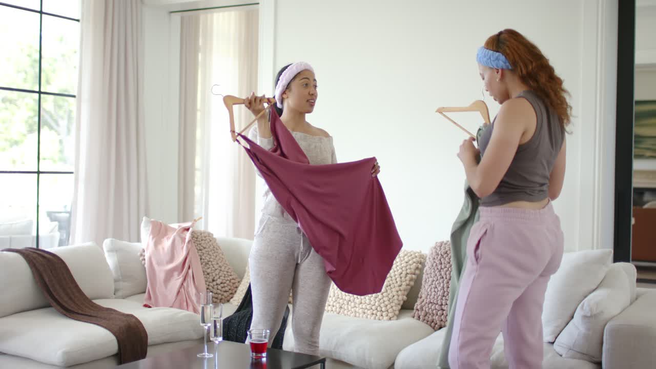 Two friends are bending to pick up, comparing maroon and green garments by sofa, choosing outfits