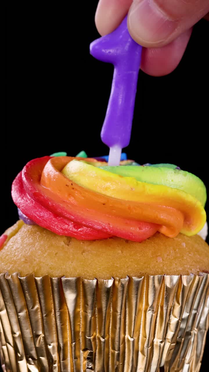 Uma mão insere uma vela roxa com o número um em um cupcake com cobertura de arco-íris e forminha dourada, sob iluminação de estúdio brilhante contra um fundo preto.