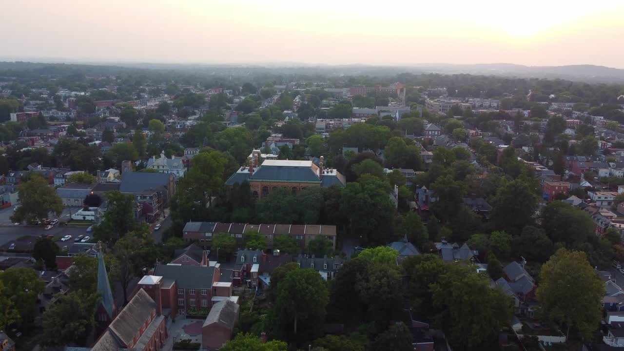 A wide drone shot over Lancaster, PA in summer, panning higher to reveal lush trees, city homes, glowing buildings, and a vibrant evening sky with distant mountains—capturing nature and urban charm.