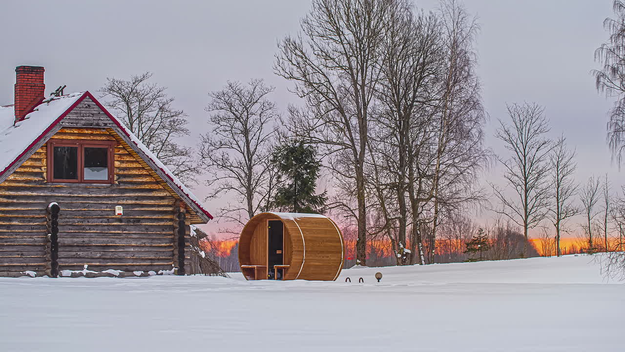 sauna de barril redondo fuera de una cabaña de troncos en la nieve en invierno con el sol saliendo en segundo plano