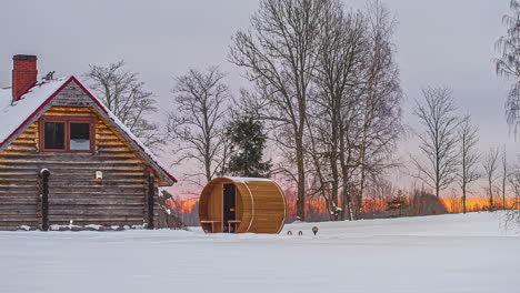 round barrel sauna outside a log cabin in snow at winter with sun rising in background