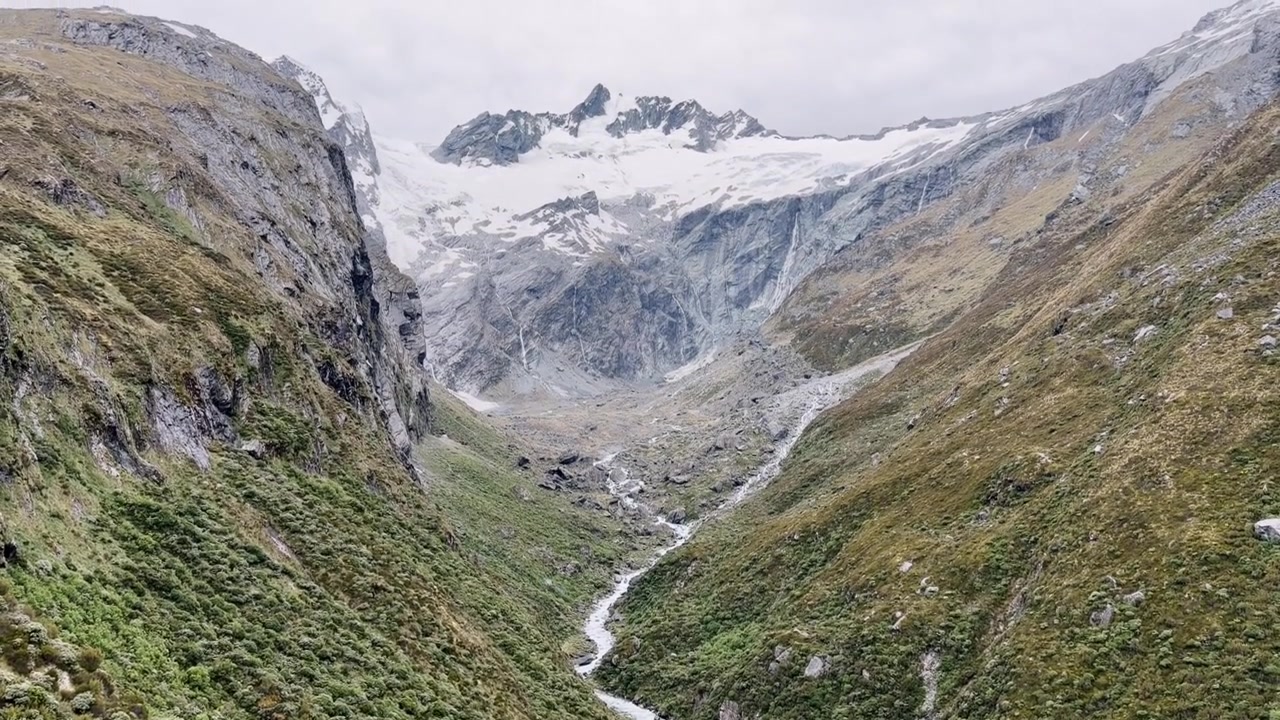 Vista panoramica di un ruscello alimentato da ghiacciaio che serpeggia attraverso una aspra valle alpina sotto una montagna innevata e un ghiacciaio nel Parco Nazionale del Monte Aspiring, Nuova Zelanda