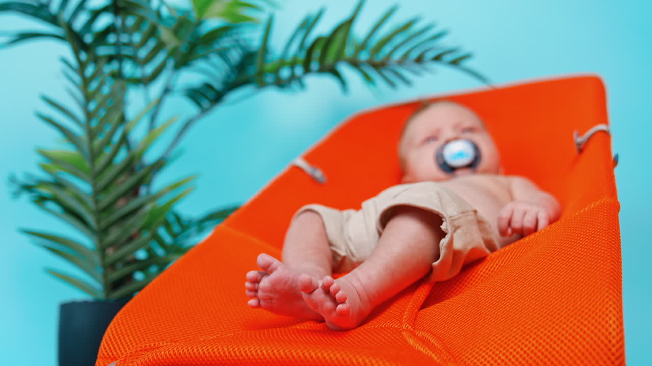Little baby boy sitting in orange chair his feet to camera. Green pot with plant standing beside. Blurred backdrop. Low angle view.