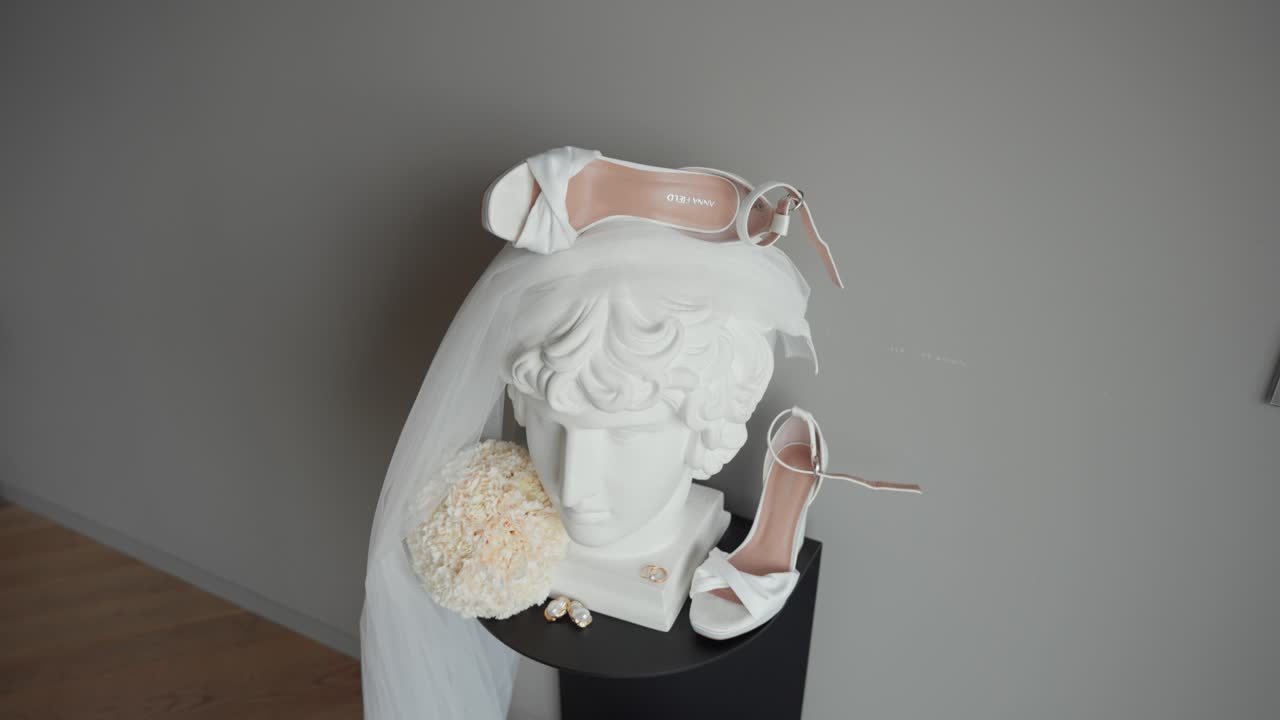 Elegant flat lay of bridal accessories featuring white heels, veil, floral bouquet, pearl earrings, and gold rings displayed on a classical bust sculpture