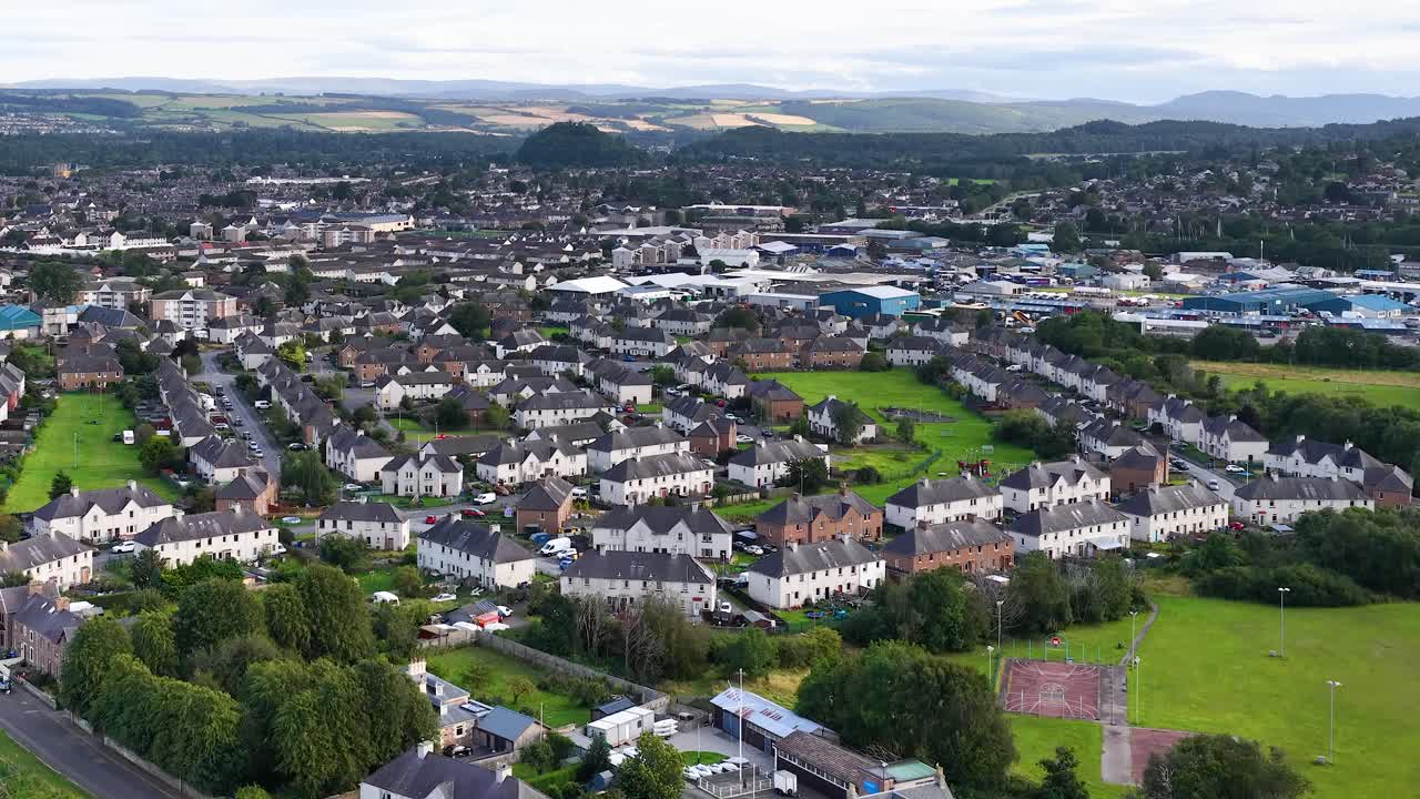 Drone glides over Dundee suburb, revealing residential rooftops, green spaces, and distant hills under daylight