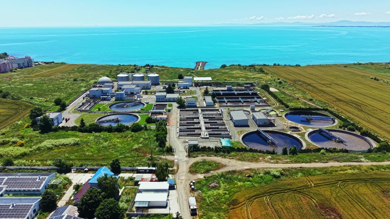 Water treatment facility located near the coastline with various large tanks and plant structures surrounded by fields and water during daytime