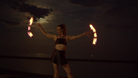 woman twirling with spinning fire fans on lakeshore beach, night exterior slowmo