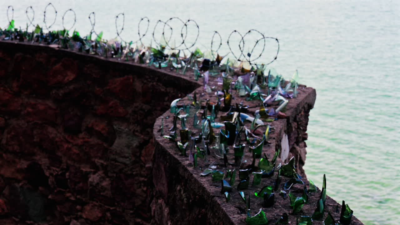 Close up of multiple broken glass shards and barbed wire loops along the edge of a brick wall with a blurred view of the sea