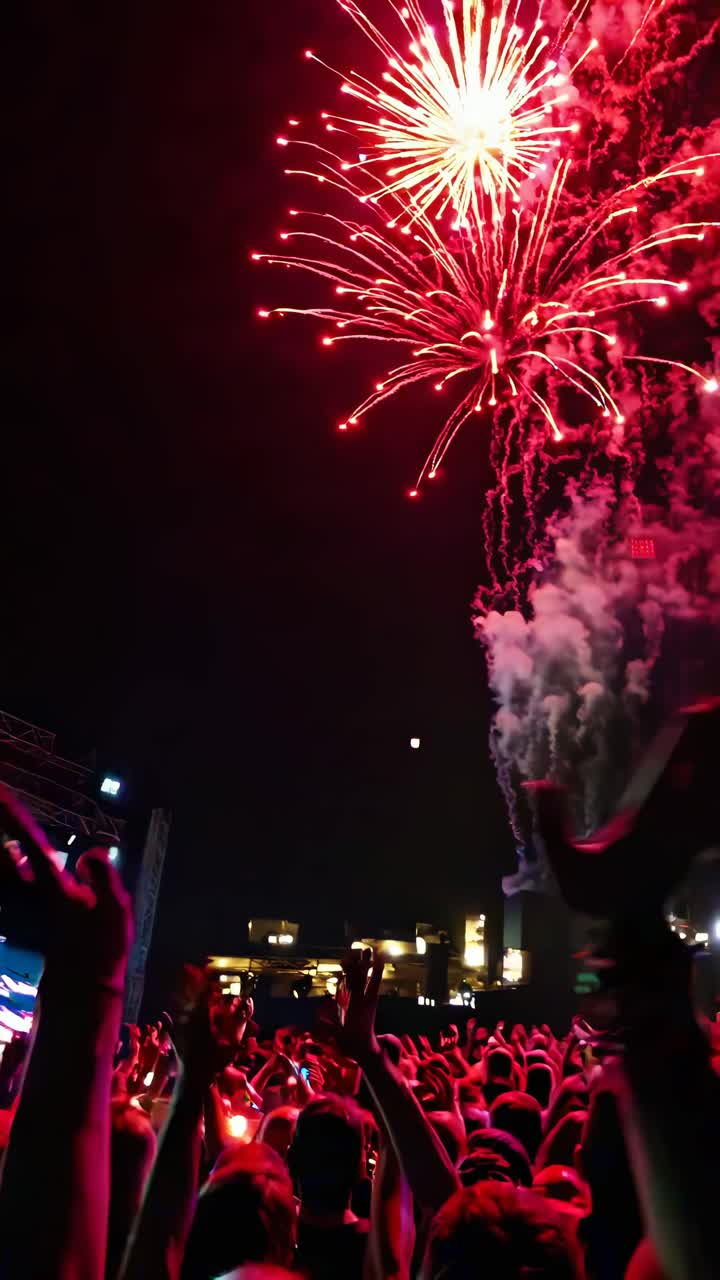 Vídeo em ângulo baixo capturando vibrantes fogos de artifício vermelhos explodindo no céu noturno acima de uma multidão animada.