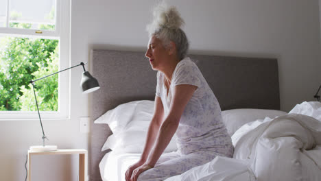 Sleepy-senior-caucasian-woman-sitting-on-the-bed-at-home