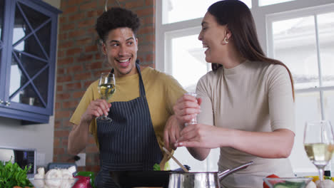 happy biracial couple cooking together and drinking wine in kitchen