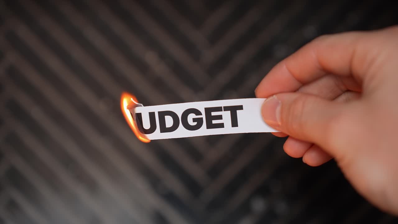 Close-up hand holds a paper strip reading “BUDGET” burning at the edge, symbolizing cutting expenses, financial planning, and breaking bad money habits with copy space