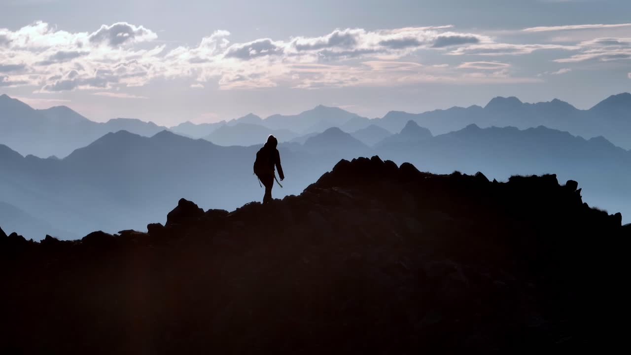 montañero femenino caminando sobre una cresta retroiluminada por el sol y otras montañas en el fondo