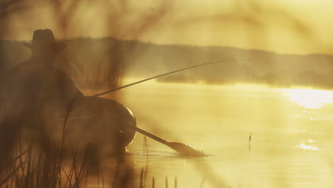 vista trasera de un anciano con sombrero en un bote pescando con una caña en el lago en una mañana nublada