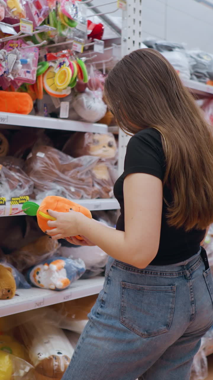 vista trasera de una mujer con una camiseta negra sosteniendo e inspeccionando un bolso de peluche naranja vibrante con detalles verdes en un centro comercial bien iluminado, examina cuidadosamente el artículo mientras navega por los estantes llenos de juguetes coloridos