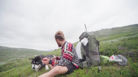 handsome man with beard eating fruit with his alaskan malamute on a grassy mountain peak