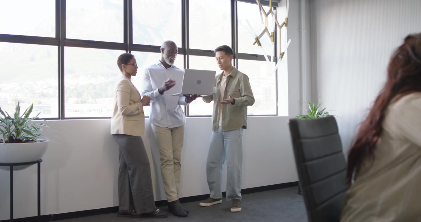 Team eterogeneo vicino alla finestra dell'ufficio, il collega al centro che apre il laptop, gli altri che si sporgono, allineandosi sul contenuto.