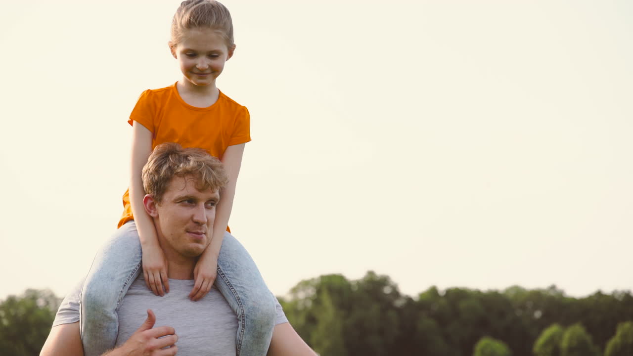 padre feliz cargando a su hija sobre los hombros en un parque mientras camina y le explica algo