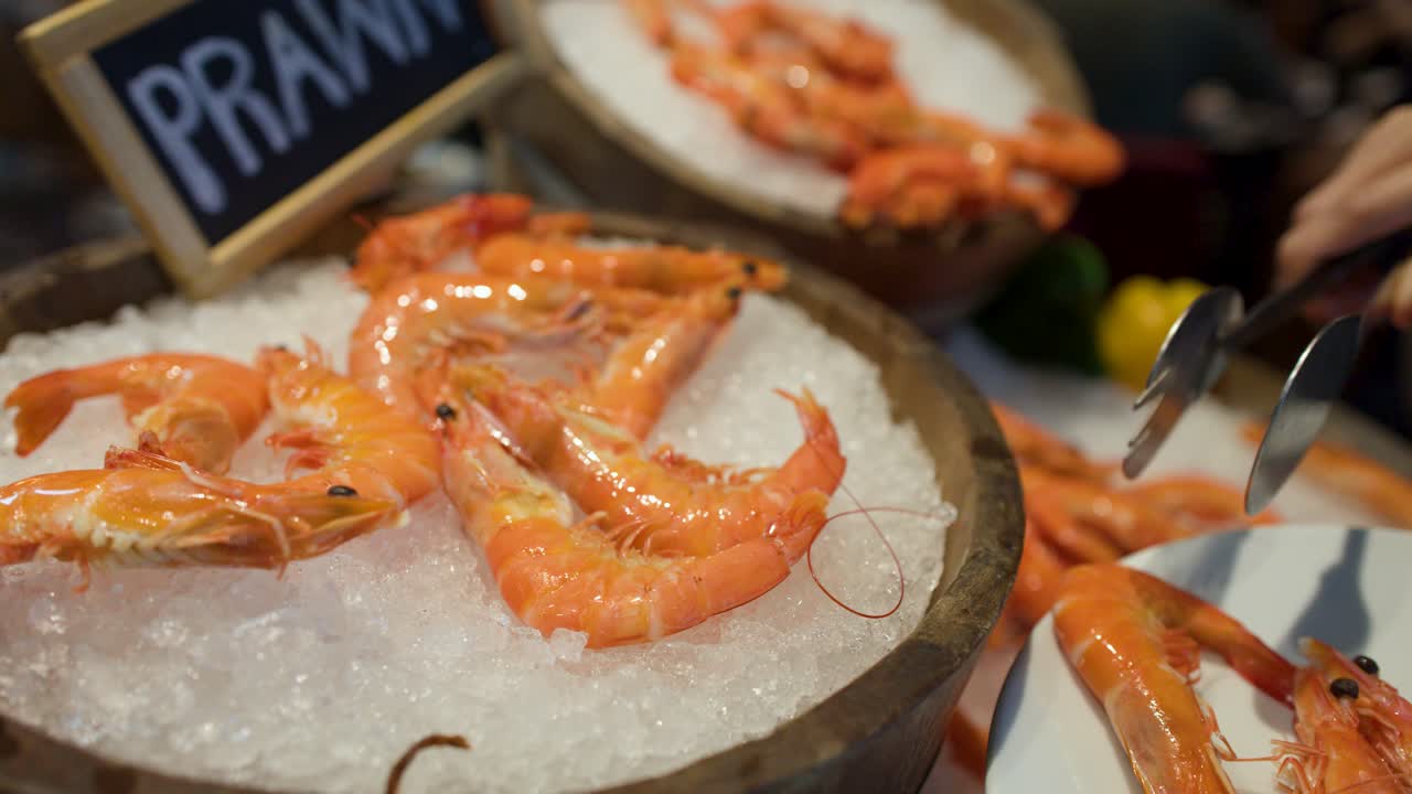 A hand uses metal tongs to select cooked prawns from a wooden bowl on ice