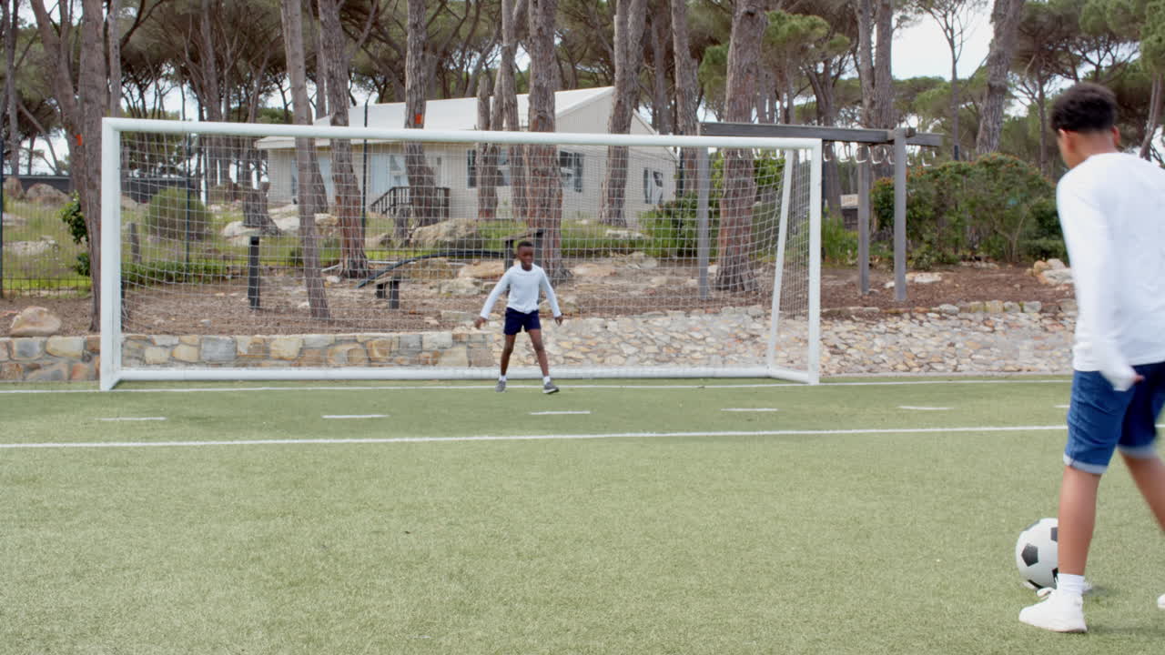 Multiracial boys playing soccer on field, one kicking ball towards goal, enjoying game