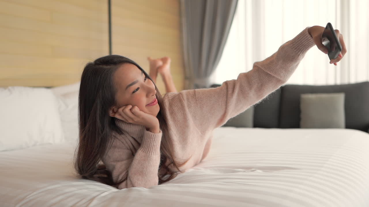 Young adult woman lying on bed taking selfie with smartphone, making video call