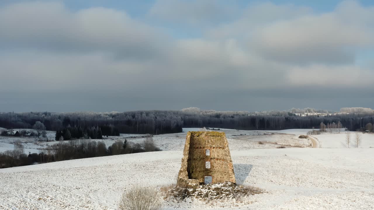 Aerial drone shot of ancient windmill structure on a snow-covered hilltop, overlooking a frozen forest in the countryside on a sunny winter day. Vast winter landscape views.