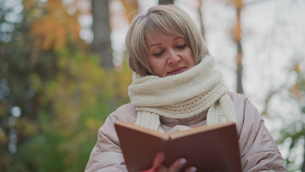 close up of woman in warm scarf and jacket focusing on open book while seated in forest setting, with soft blurry autumn trees in background creating serene and peaceful outdoor reading atmosphere
