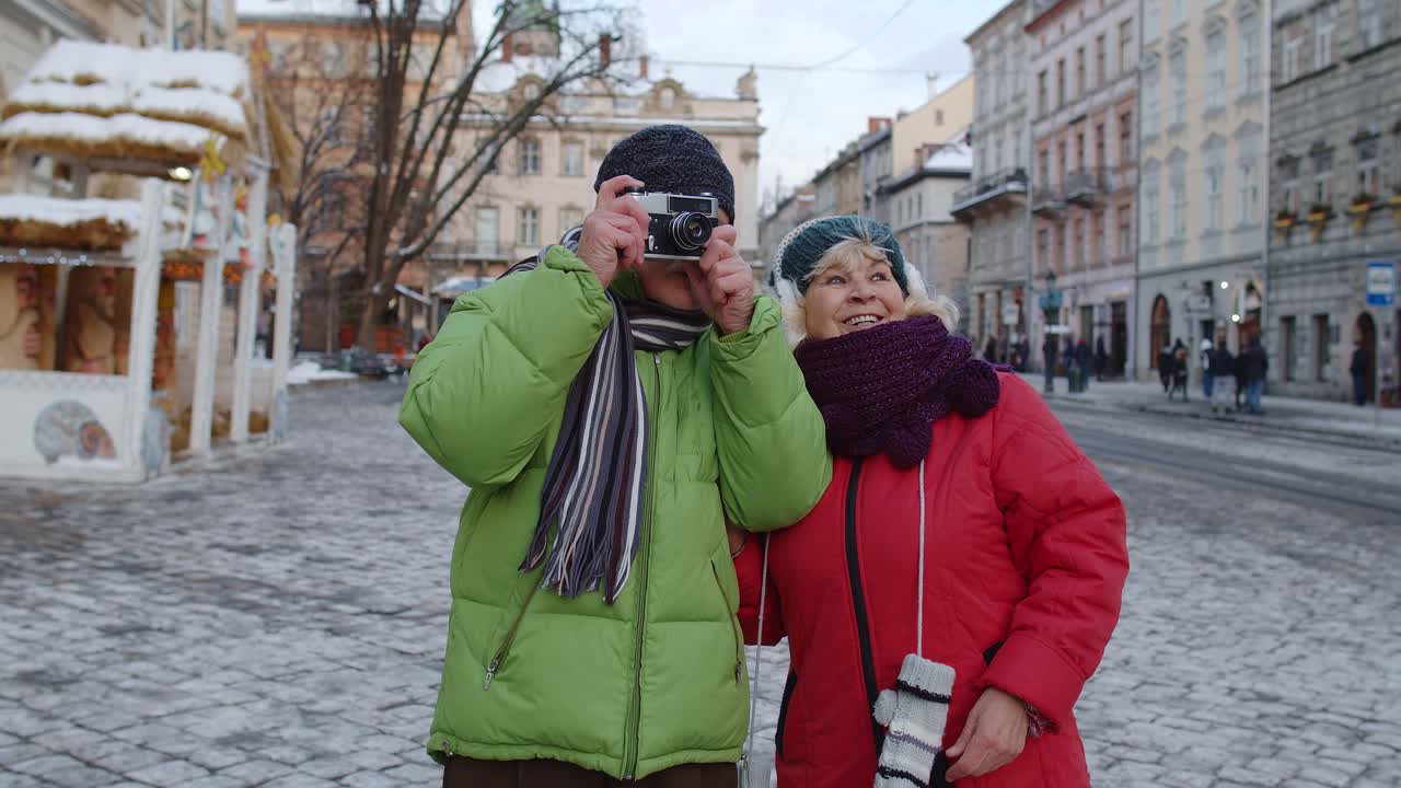 Coppia di anziani turisti, marito e moglie, che scattano foto con una macchina fotografica retrò, passeggiando per una strada cittadina invernale.