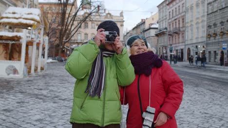 Senior-wife-husband-tourists-taking-photo-pictures-on-retro-camera,-walking-on-winter-city-street