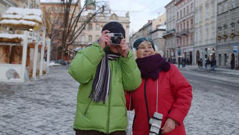 senior wife husband tourists taking photo pictures on retro camera, walking on winter city street