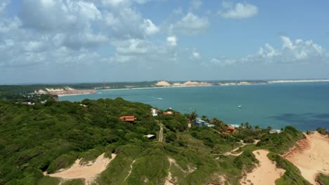 Rising-aerial-drone-shot-of-large-sand-dunes-revealing-the-tropical-famous-tourist-beach-town-of-Pipa,-Brazil-in-Rio-Grande-do-Norte-with-boats-in-the-distance-in-the-turquoise-ocean-on-a-summer-day