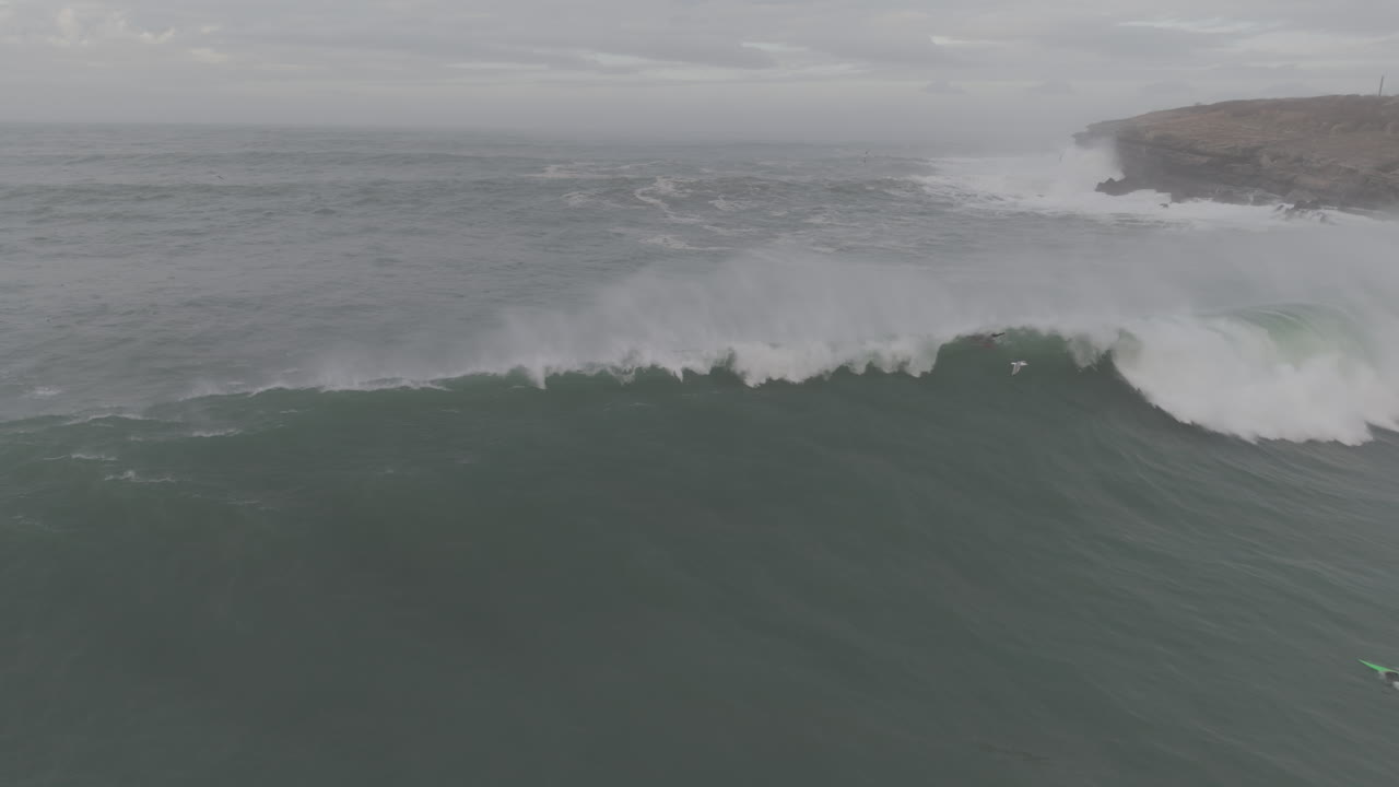 Aerial View of Surfers Riding a Massive Ocean Wave Near a Rocky Coastline