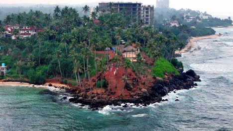 high-angle telephoto captures sri lanka's coast visitors explore a red-earth headland with green palms under hazy skies, beside dark rocks, waves, and a curving beach