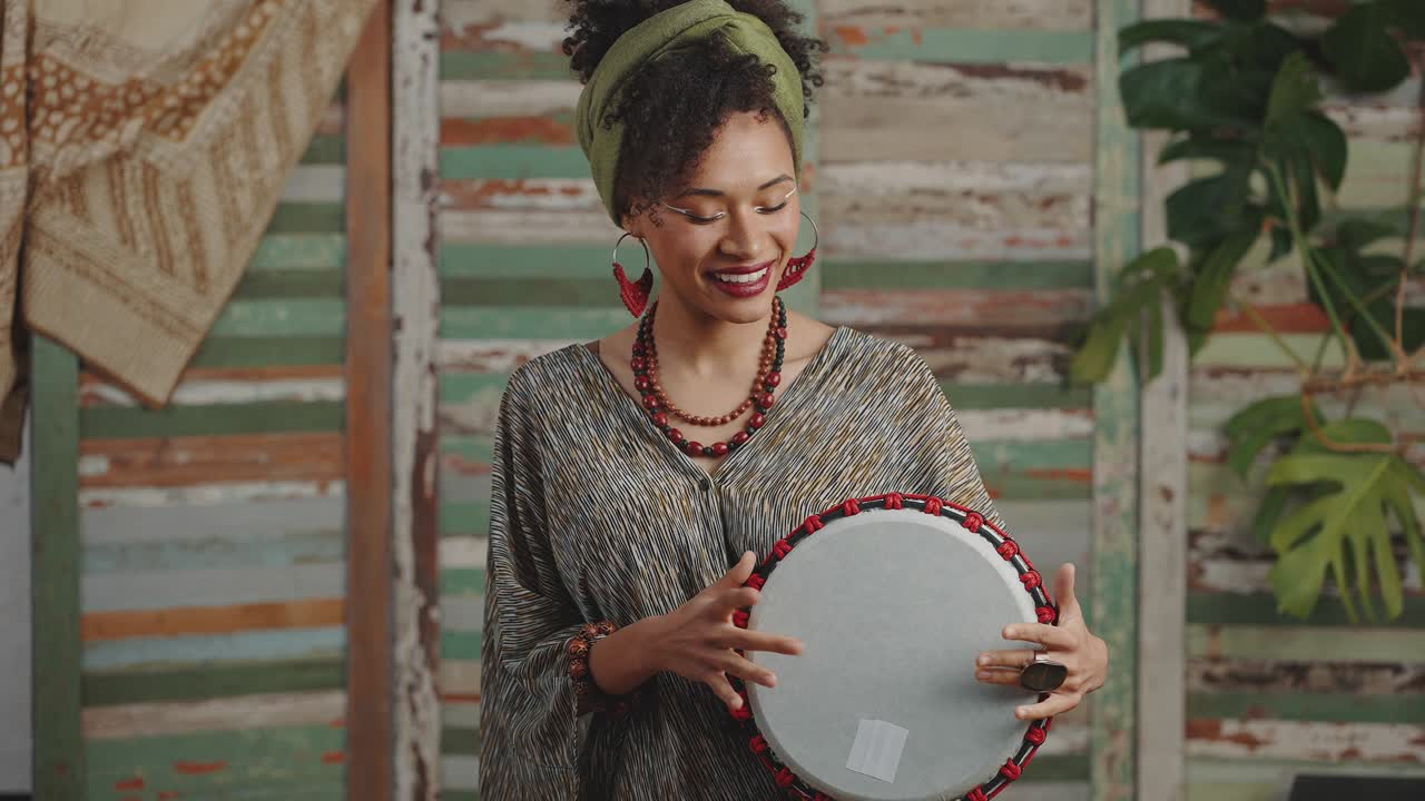joven feliz y elegante mujer afroamericana tocando el djembe, tocando la batería y sonriendo a la cámara