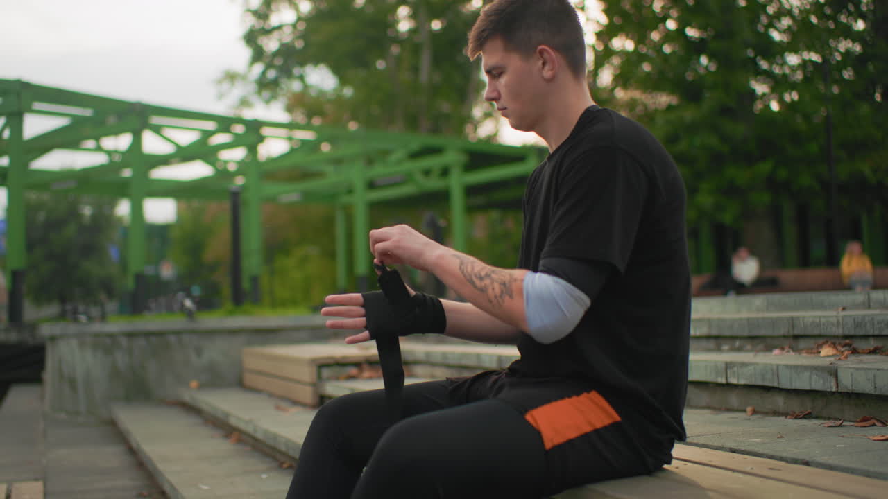 Uomo bianco che pratica la terapia del bastone da passeggio sui gradini di una panchina vicino a uno stagno, esercizio di riabilitazione incentrato su equilibrio e postura, routine guidata, presa tattile, tatuaggi visibili, abbigliamento sportivo casual, tardi