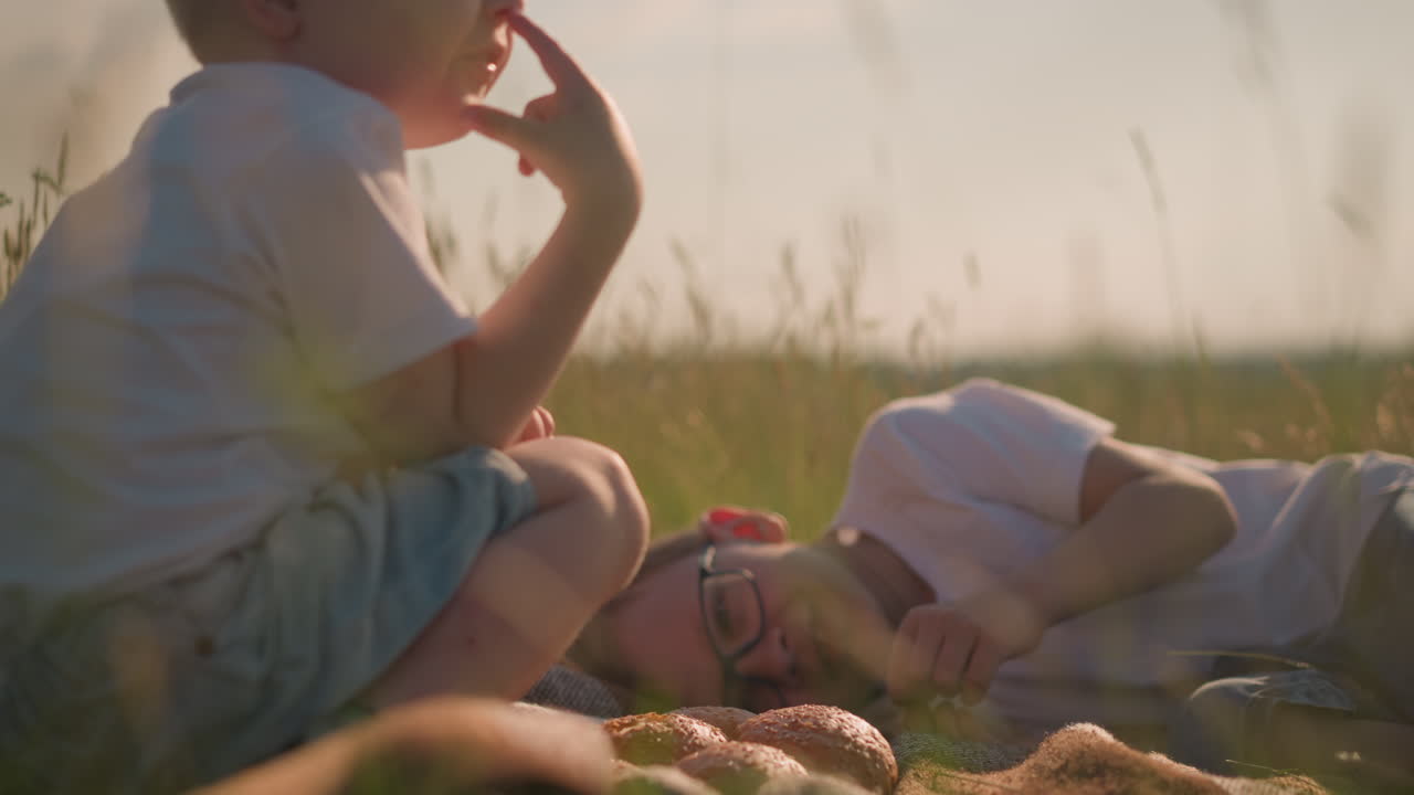 A boy wearing glasses and a white shirt lies on a scarf in a grassy field, gently touching a piece of bread. Beside him, his younger brother, also in a white shirt, touches his nose in a thoughtful