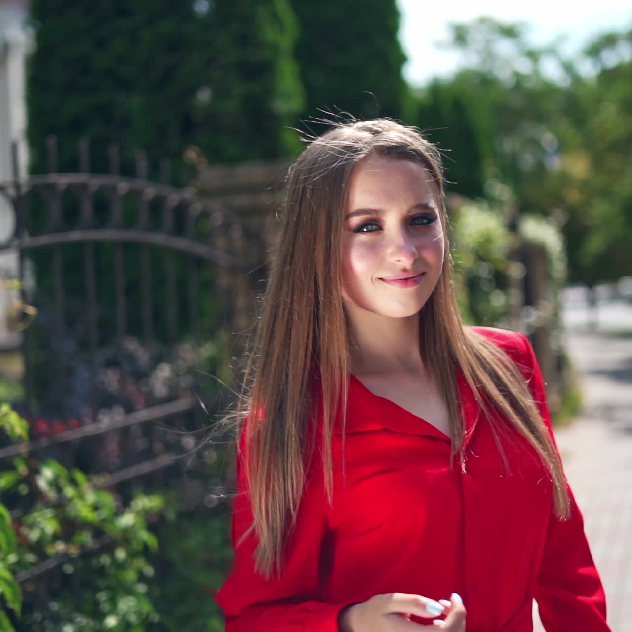 Lovely girl in red dress. Beautiful young model is standing outdoors and posing on camera. Portrait of a pretty woman on the green trees background.