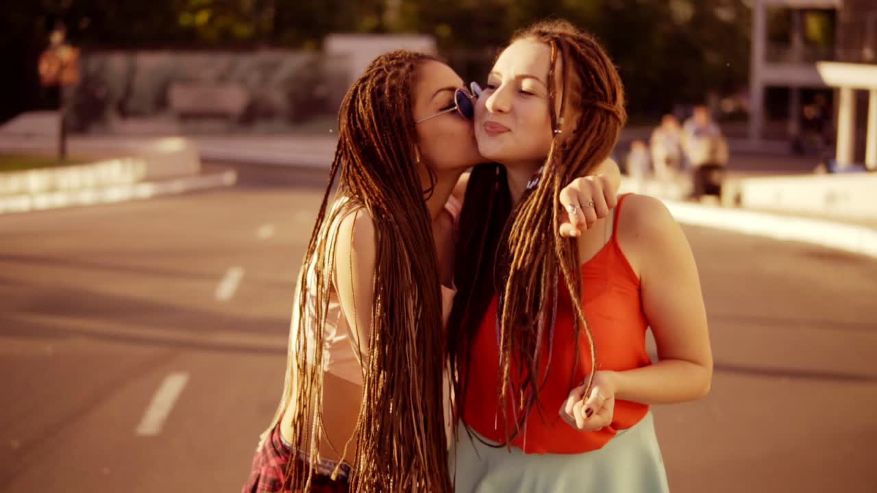 Two happy women with dreads walking on the empty road and talking in summer. Two hipster girls laughing, hugging and kissing