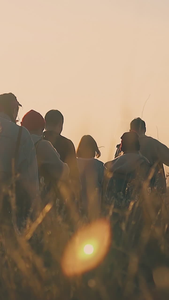 group of young tourists in warm clothes walk on meadow with dry grass in early autumn morning at back sunlight slow motion