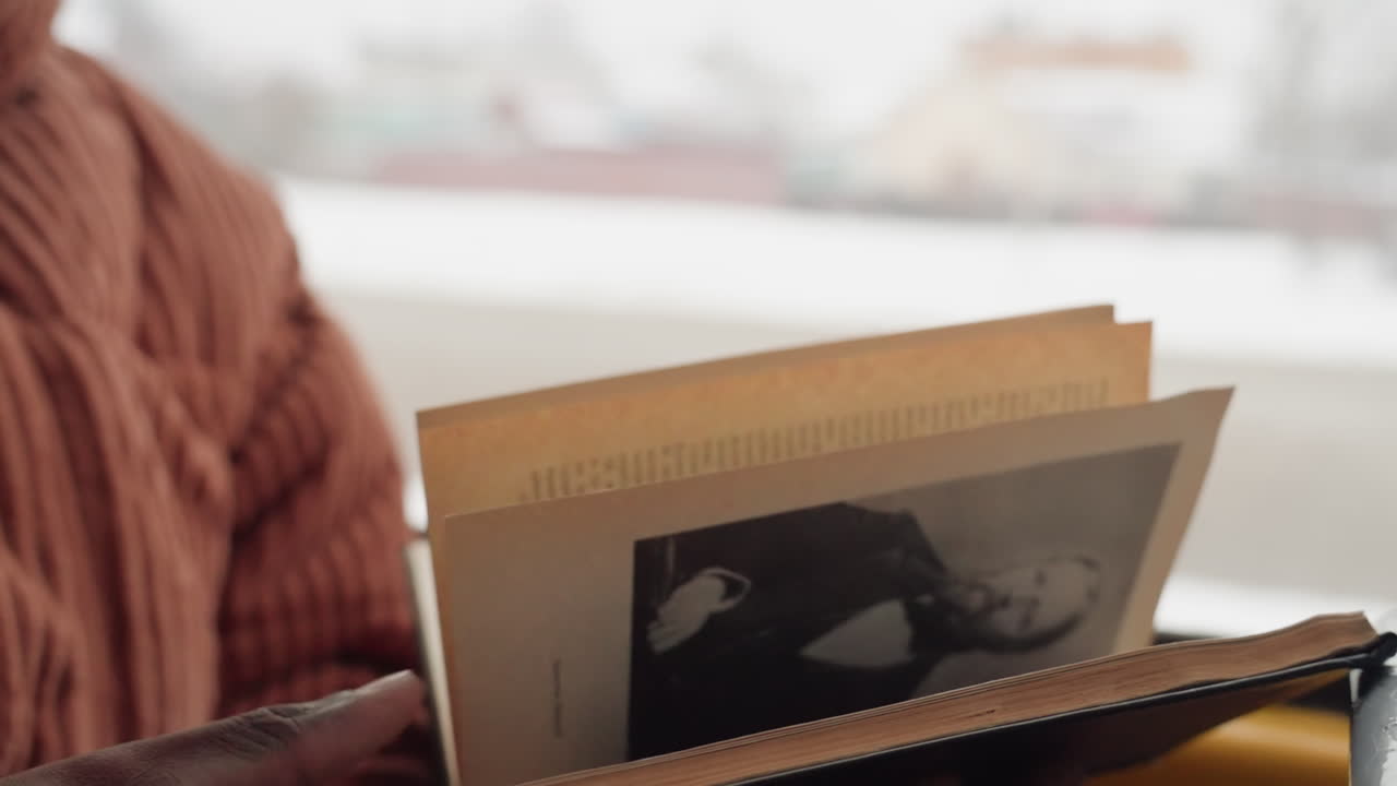 Pendolare che legge un libro vicino al finestrino, tiene un taccuino nero con copertina rigida e fotografia seppia, dita che girano le pagine, paesaggio urbano innevato visibile attraverso il finestrino ghiacciato del treno, caldo maglione di lana, atmosfera tranquilla.