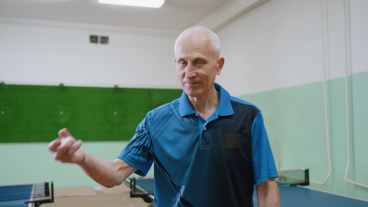 Old man in blue sports shirt bouncing tennis ball on indoor floor near table tennis tables, demonstrating coordination, focus, balance, agility, active lifestyle, strength, and healthy aging