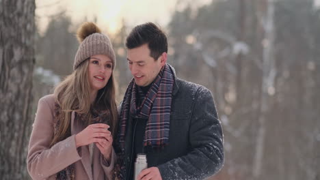 beautiful and stylish couple man and woman hipsters in a coat and scarf drink tea from a thermos in the winter forest after a walk. love story