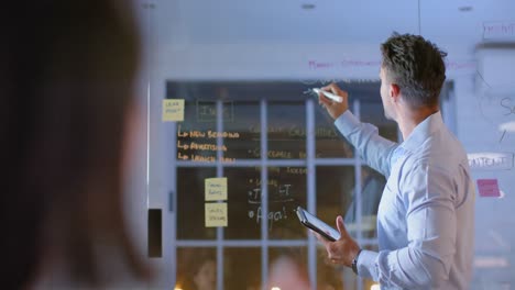 Biracial-businessman-using-tablet-and-glass-wall-presenting-to-colleague-at-night-in-office