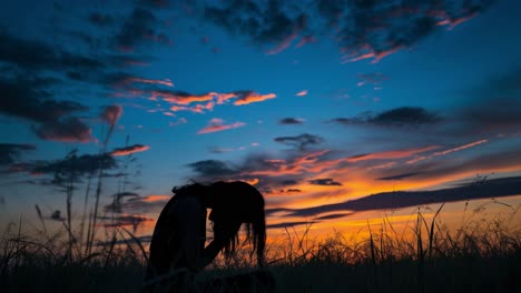 silhouetting a young woman praying with joined hands in a tall grass field at sunset, showcasing a serene sky of orange, pink, and blue hues