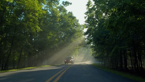 Blue-car-driving-on-road-near-foggy-forest-with-sun-beaming-through-canopy-in-the-morning