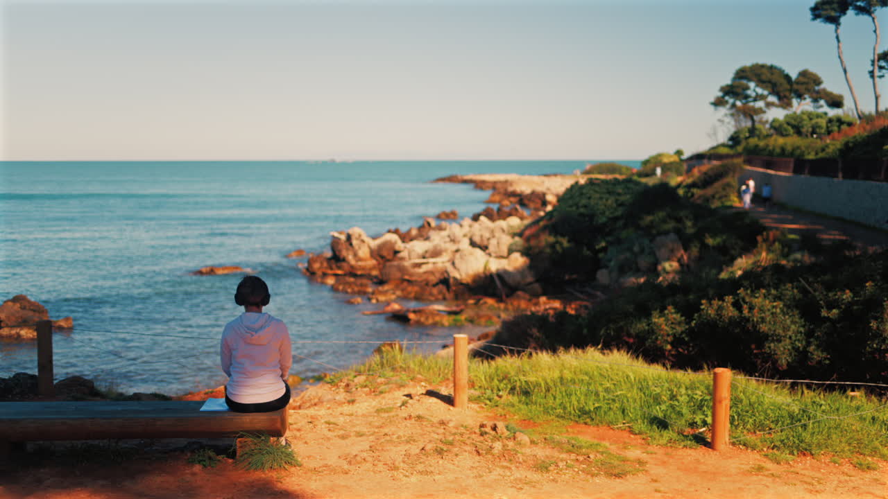 Woman with headphones on her head sitting on a wooden bench with a view of the sea
